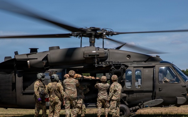 A group of U.S. Army soldiers are loading or unloading a casualty onto a UH-60 Black Hawk helicopter on a grassy field. The helicopter's rotors are spinning, indicating it is either landing or preparing for takeoff. This appears to be a medical evacuation (MEDEVAC) exercise or operation, evidenced by the red cross marking on the aircraft and the coordinated team effort.