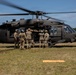 A group of U.S. Army soldiers are loading or unloading a casualty onto a UH-60 Black Hawk helicopter on a grassy field. The helicopter's rotors are spinning, indicating it is either landing or preparing for takeoff. This appears to be a medical evacuation (MEDEVAC) exercise or operation, evidenced by the red cross marking on the aircraft and the coordinated team effort.