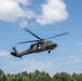 A military medical evacuation (MEDEVAC) helicopter, identifiable as a UH-60 Black Hawk, is captured in flight against a partly cloudy blue sky. The aircraft bears a visible red cross emblem on its fuselage, indicating its medical/humanitarian role. It is flying at low altitude above a treeline in what appears to be a daytime operational or training scenario.