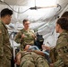 A group of U.S. Army medical personnel are gathered inside a military medical tent around a patient lying on a stretcher with an airway tube in place. A senior officer, likely a medical officer based on rank insignia, appears to be instructing or briefing the team. The setting suggests a field medical training exercise or forward surgical team operation.