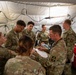 A group of U.S. Army medical personnel are gathered inside a military field hospital tent, attending to a patient lying on a treatment table. The scene appears to be a medical training exercise or active triage situation, with multiple soldiers in OCP (Operational Camouflage Pattern) uniforms surrounding the patient. Medical equipment, supplies, and a red tool cart with medical supplies are visible in the background.