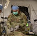 Two U.S. Army medical personnel in military camouflage uniforms are performing a medical procedure inside what appears to be a military field hospital tent. The central figure, identified by name tape as 'BARNHILL' with captain rank insignia, is wearing surgical PPE including gloves, mask, and scrub cap while working on a patient. Medical equipment including IV stands, monitoring devices, and a crash cart are visible in the background.