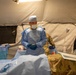 A medical professional in full surgical attire is performing or preparing for a medical procedure on a patient inside a military field hospital tent. The setting appears to be a forward operating or expeditionary medical facility, indicated by the tent structure and military uniforms visible on both the patient and an assisting personnel. The scene is well-lit by an overhead surgical lamp.