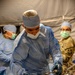 A medical professional in full surgical attire is performing or preparing a procedure in what appears to be a military field surgical environment. At least two other personnel are visible in the background, one wearing military camouflage (OCP pattern) with a visible medical unit patch. The setting appears to be inside a military field hospital tent.