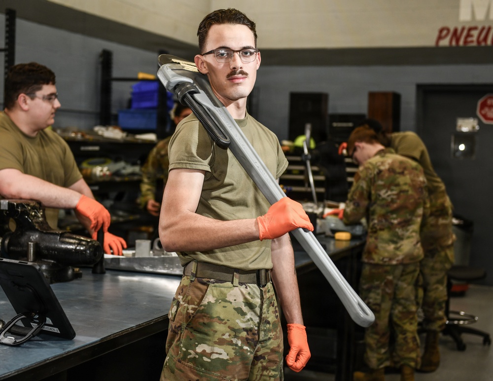 A military service member in OCP (Operational Camouflage Pattern) uniform stands in the foreground of a maintenance workshop, holding a large pipe wrench over his shoulder while wearing orange protective gloves. He is looking directly at the camera with a confident expression. Several other uniformed personnel are visible in the background working at workbenches in what appears to be a military maintenance facility.