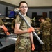 A military service member in OCP (Operational Camouflage Pattern) uniform stands in the foreground of a maintenance workshop, holding a large pipe wrench over his shoulder while wearing orange protective gloves. He is looking directly at the camera with a confident expression. Several other uniformed personnel are visible in the background working at workbenches in what appears to be a military maintenance facility.