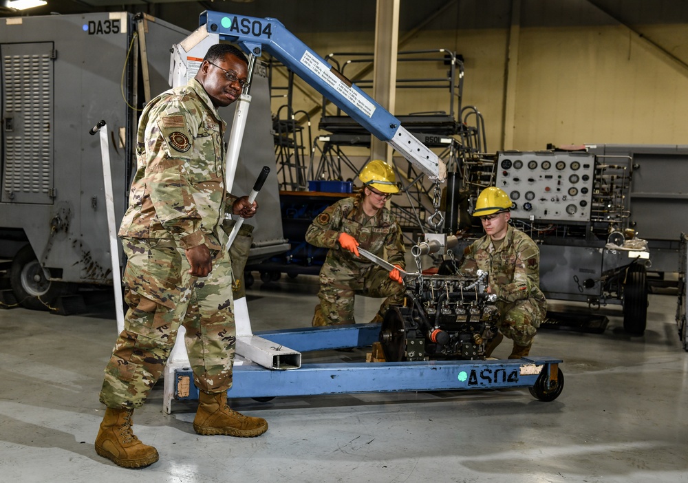Three military personnel in OCP (Operational Camouflage Pattern) uniforms are working in what appears to be a maintenance hangar or workshop. One service member is operating a blue engine hoist/crane cart labeled 'AS04', while two others wearing yellow hard hats work on what appears to be an engine or mechanical component. The setting contains various ground support equipment and machinery in the background.