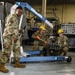 Three military personnel in OCP (Operational Camouflage Pattern) uniforms are working in what appears to be a maintenance hangar or workshop. One service member is operating a blue engine hoist/crane cart labeled 'AS04', while two others wearing yellow hard hats work on what appears to be an engine or mechanical component. The setting contains various ground support equipment and machinery in the background.