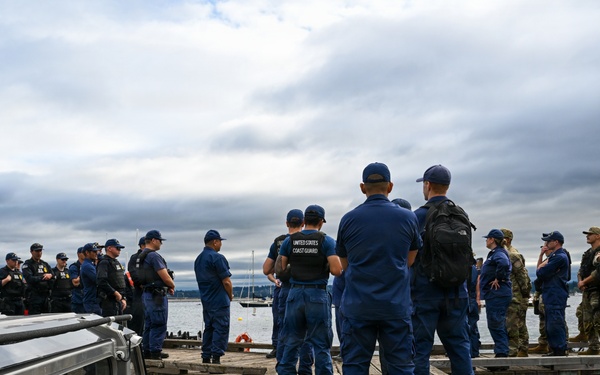 A group of United States Coast Guard personnel and military soldiers are assembled on a wooden dock near watercraft. The gathering appears to be a briefing or pre-operation muster, with personnel in both Coast Guard blue uniforms and military camouflage attire. The scene is set on a waterfront with boats docked on either side of the pier.