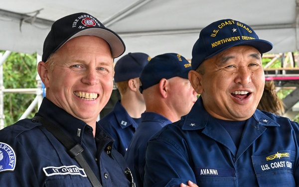 Two uniformed public safety officials are posing together and smiling under a tent or canopy structure. The man on the left wears a Seattle Fire Department uniform with the name tag 'LT. O'BRIEN' and a radio, while the man on the right wears a U.S. Coast Guard Northwest District uniform with the name tag 'AVANNI'. Additional Coast Guard personnel are visible in the background.
