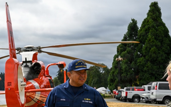 Three military personnel are conversing outdoors near a U.S. Coast Guard helicopter with distinctive orange and white coloring. The center individual is wearing a U.S. Coast Guard uniform with a 'Northwest District' cap and name tag reading 'AVANNI,' while the other two wear olive/green flight suits with American flag patches. The setting appears to be a grassy area near a body of water, likely during a public event or operational briefing.
