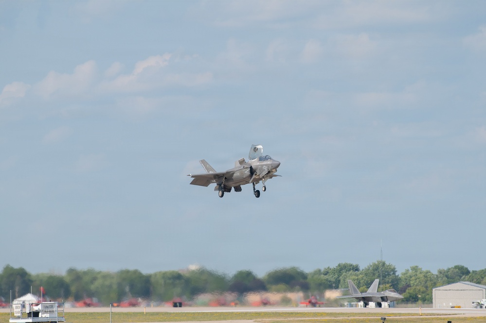 A military fighter jet, identifiable as an F-35 Lightning II, is captured in the process of taking off or landing at a military airfield with landing gear deployed. A second aircraft, appearing to be an F-22 Raptor, is visible on the tarmac in the background. The scene appears to be an air show or active military base, with spectator areas and support structures visible in the background.