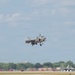 A military fighter jet, identifiable as an F-35 Lightning II, is captured in the process of taking off or landing at a military airfield with landing gear deployed. A second aircraft, appearing to be an F-22 Raptor, is visible on the tarmac in the background. The scene appears to be an air show or active military base, with spectator areas and support structures visible in the background.