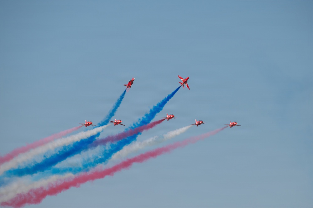 The image shows a formation of red jet aircraft, identifiable as the RAF Red Arrows aerobatic display team, performing a maneuver in a clear blue sky. The aircraft are emitting red, white, and blue smoke trails, which are characteristic of their signature displays. The formation appears to be in the process of a split or crossover maneuver.
