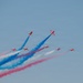 The image shows a formation of red jet aircraft, identifiable as the RAF Red Arrows aerobatic display team, performing a maneuver in a clear blue sky. The aircraft are emitting red, white, and blue smoke trails, which are characteristic of their signature displays. The formation appears to be in the process of a split or crossover maneuver.