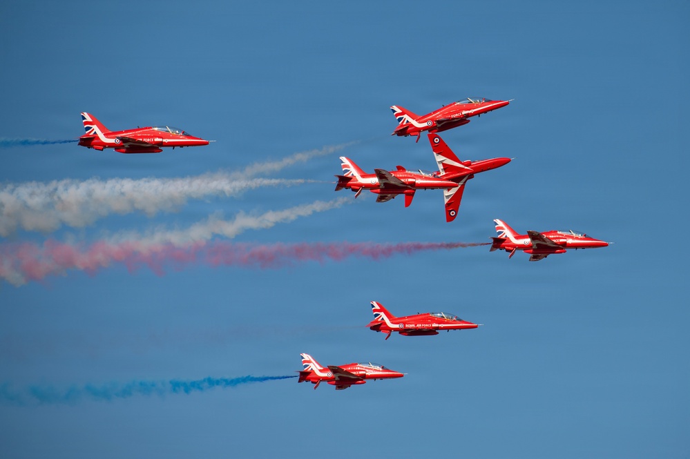 The image shows the RAF Red Arrows aerobatic display team performing a formation maneuver against a clear blue sky. Seven BAE Hawk T1 jet aircraft in their distinctive red livery are captured mid-display, with colored smoke trails in red, white, and blue visible behind several aircraft. The jets appear to be executing a break or split formation maneuver.