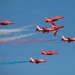 The image shows the RAF Red Arrows aerobatic display team performing a formation maneuver against a clear blue sky. Seven BAE Hawk T1 jet aircraft in their distinctive red livery are captured mid-display, with colored smoke trails in red, white, and blue visible behind several aircraft. The jets appear to be executing a break or split formation maneuver.