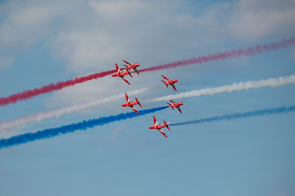 The image shows a formation of red jet aircraft performing an aerobatic display, trailing red, white, and blue smoke streams across a blue sky. The aircraft appear to be the British Royal Air Force Red Arrows aerobatic display team flying in a diamond-split formation. The colorful smoke trails create a striking visual pattern consistent with a patriotic air show performance.