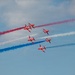 The image shows a formation of red jet aircraft performing an aerobatic display, trailing red, white, and blue smoke streams across a blue sky. The aircraft appear to be the British Royal Air Force Red Arrows aerobatic display team flying in a diamond-split formation. The colorful smoke trails create a striking visual pattern consistent with a patriotic air show performance.