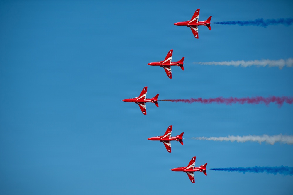 Five red jet aircraft fly in a diagonal echelon formation against a clear blue sky. The aircraft are emitting colored smoke trails in red, white, and blue, consistent with a precision aerobatic display team performance. The formation and livery are characteristic of the British Royal Air Force Red Arrows display team flying BAE Hawk jets.