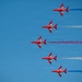 Five red jet aircraft fly in a diagonal echelon formation against a clear blue sky. The aircraft are emitting colored smoke trails in red, white, and blue, consistent with a precision aerobatic display team performance. The formation and livery are characteristic of the British Royal Air Force Red Arrows display team flying BAE Hawk jets.