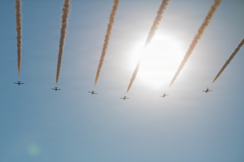 Seven aircraft are flying in a diagonal formation across a clear blue sky, each emitting smoke trails behind them. The formation appears to be part of an aerobatic or airshow display, with the sun creating a bright glare in the upper right portion of the image. The smoke trails are parallel and evenly spaced, indicating precise coordinated flight.