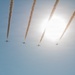 Seven aircraft are flying in a diagonal formation across a clear blue sky, each emitting smoke trails behind them. The formation appears to be part of an aerobatic or airshow display, with the sun creating a bright glare in the upper right portion of the image. The smoke trails are parallel and evenly spaced, indicating precise coordinated flight.