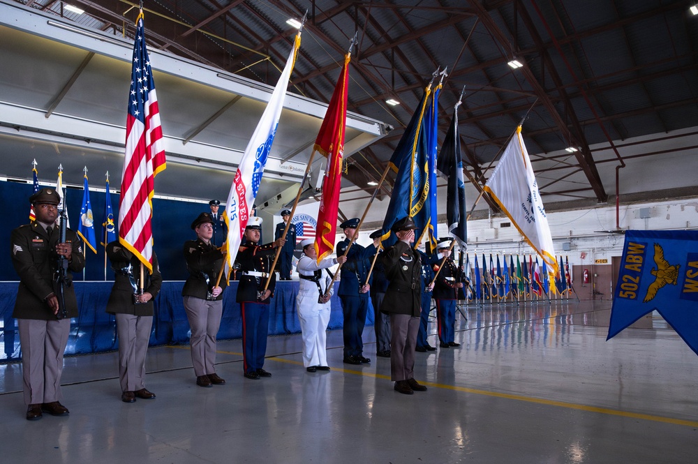 A joint military color guard ceremony is taking place inside what appears to be an aircraft hangar or large military facility. Personnel from multiple branches of the U.S. Armed Forces are presenting flags, including the American flag and various service and unit colors. A banner for the 502nd Air Base Wing (502 ABW) is visible on the right side of the image.
