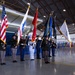 A joint military color guard ceremony is taking place inside what appears to be an aircraft hangar or large military facility. Personnel from multiple branches of the U.S. Armed Forces are presenting flags, including the American flag and various service and unit colors. A banner for the 502nd Air Base Wing (502 ABW) is visible on the right side of the image.
