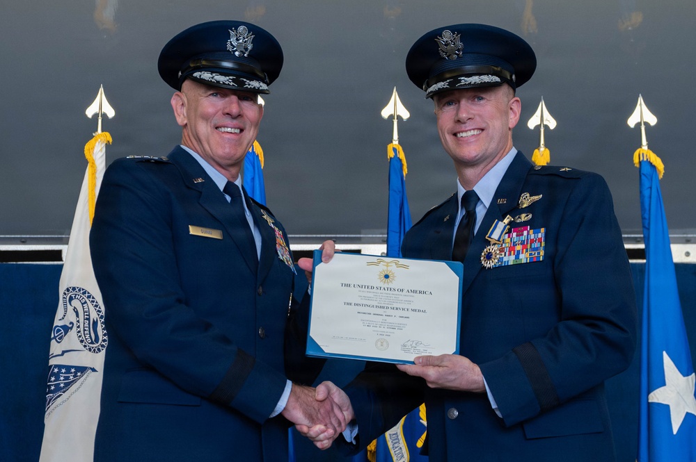 Two United States Air Force officers in dress blue uniforms are shaking hands while jointly holding a Distinguished Service Medal certificate. The ceremony appears to be a formal military awards presentation, with flags and ceremonial elements visible in the background. Both officers are smiling, suggesting a positive and celebratory occasion.
