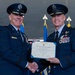 Two United States Air Force officers in dress blue uniforms are shaking hands while jointly holding a Distinguished Service Medal certificate. The ceremony appears to be a formal military awards presentation, with flags and ceremonial elements visible in the background. Both officers are smiling, suggesting a positive and celebratory occasion.