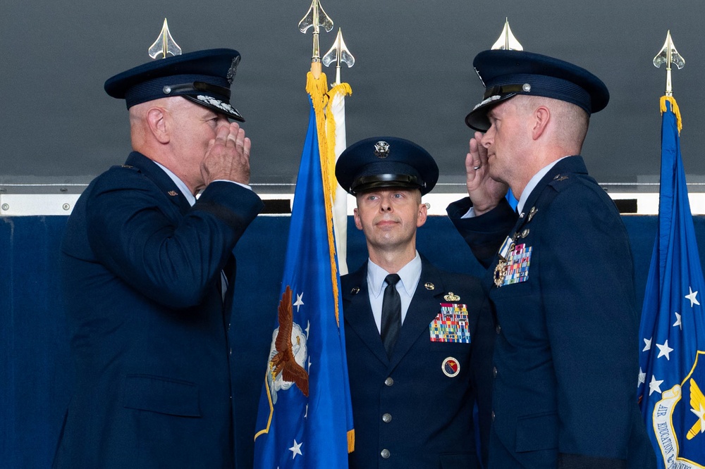 A formal U.S. Air Force military ceremony is taking place, likely a change of command. Two senior officers in dress uniforms are exchanging salutes while a third officer stands at attention holding a guidon or organizational flag in the center. Multiple Air Force flags and ceremonial standards are visible in the background.