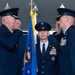 A formal U.S. Air Force military ceremony is taking place, likely a change of command. Two senior officers in dress uniforms are exchanging salutes while a third officer stands at attention holding a guidon or organizational flag in the center. Multiple Air Force flags and ceremonial standards are visible in the background.