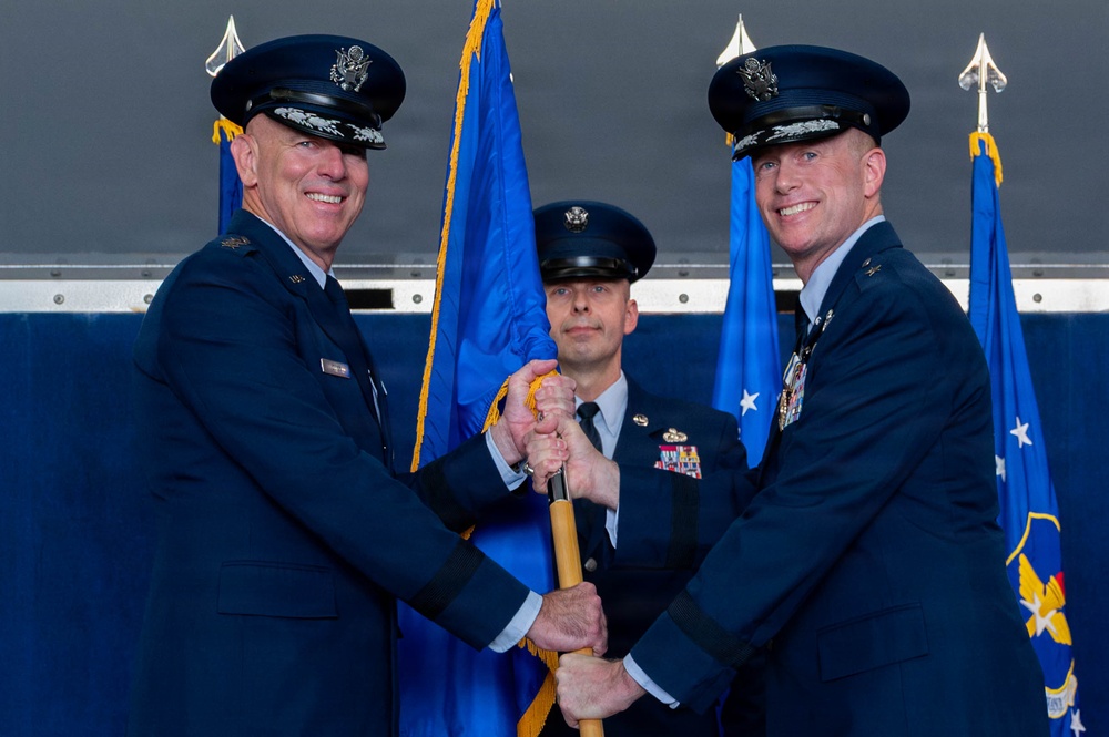 A formal United States Air Force change of command ceremony is taking place, with two senior officers exchanging a guidon (ceremonial flag staff) while a third officer stands at attention in the background. The two principal officers are wearing service dress uniforms with general officer rank insignia, medals, and service ribbons. Blue Air Force flags with gold fringe are prominently displayed in the background.