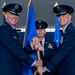 A formal United States Air Force change of command ceremony is taking place, with two senior officers exchanging a guidon (ceremonial flag staff) while a third officer stands at attention in the background. The two principal officers are wearing service dress uniforms with general officer rank insignia, medals, and service ribbons. Blue Air Force flags with gold fringe are prominently displayed in the background.