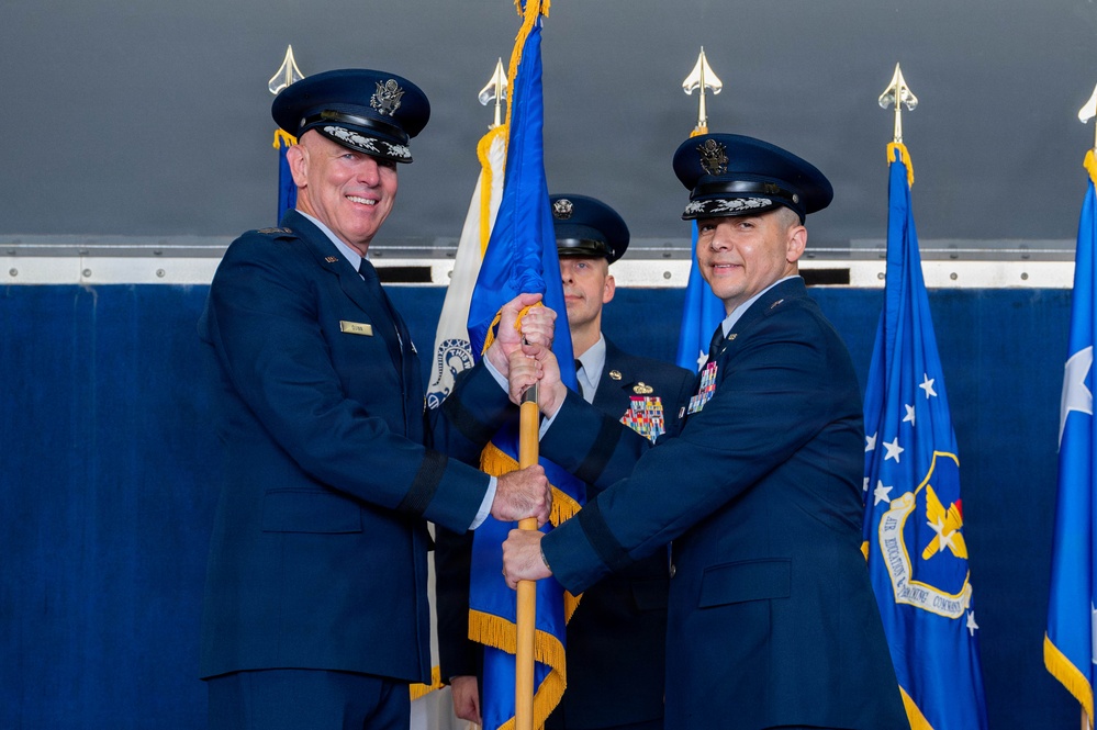 Two senior United States Air Force officers are participating in a formal change of command ceremony, passing a guidon (unit flag) between them. The officer on the left, whose name tag reads 'RYAN', appears to be a General based on his rank insignia, while the officer on the right is also a senior officer with numerous service ribbons. A third uniformed airman stands in the background holding additional flags.