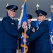 Two senior United States Air Force officers are participating in a formal change of command ceremony, passing a guidon (unit flag) between them. The officer on the left, whose name tag reads 'RYAN', appears to be a General based on his rank insignia, while the officer on the right is also a senior officer with numerous service ribbons. A third uniformed airman stands in the background holding additional flags.