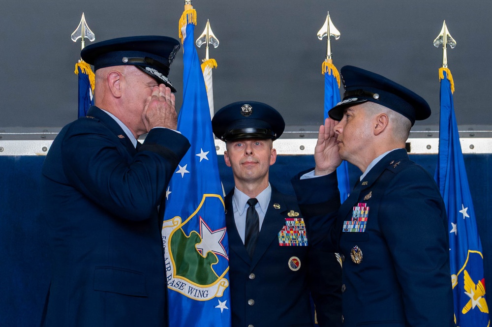 Three U.S. Air Force officers are participating in a formal military ceremony, likely a change of command. Two senior officers are exchanging salutes while a third stands at attention between them. Blue Air Force flags and a unit guidon bearing 'Air Base Wing' insignia are visible in the background.