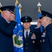 Three U.S. Air Force officers are participating in a formal military ceremony, likely a change of command. Two senior officers are exchanging salutes while a third stands at attention between them. Blue Air Force flags and a unit guidon bearing 'Air Base Wing' insignia are visible in the background.