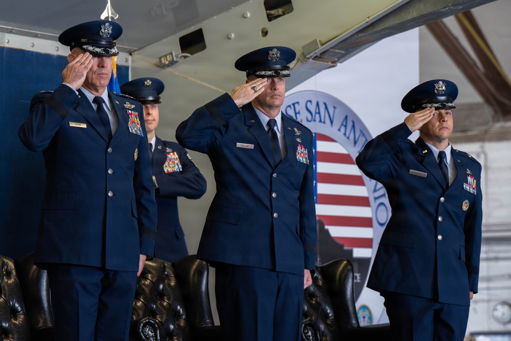 Four United States Air Force officers in dress blue uniforms are rendering a salute during a formal military ceremony. The event appears to be taking place at Joint Base San Antonio, as indicated by the partially visible banner in the background. The setting is likely a change of command or official military function held inside a hangar.