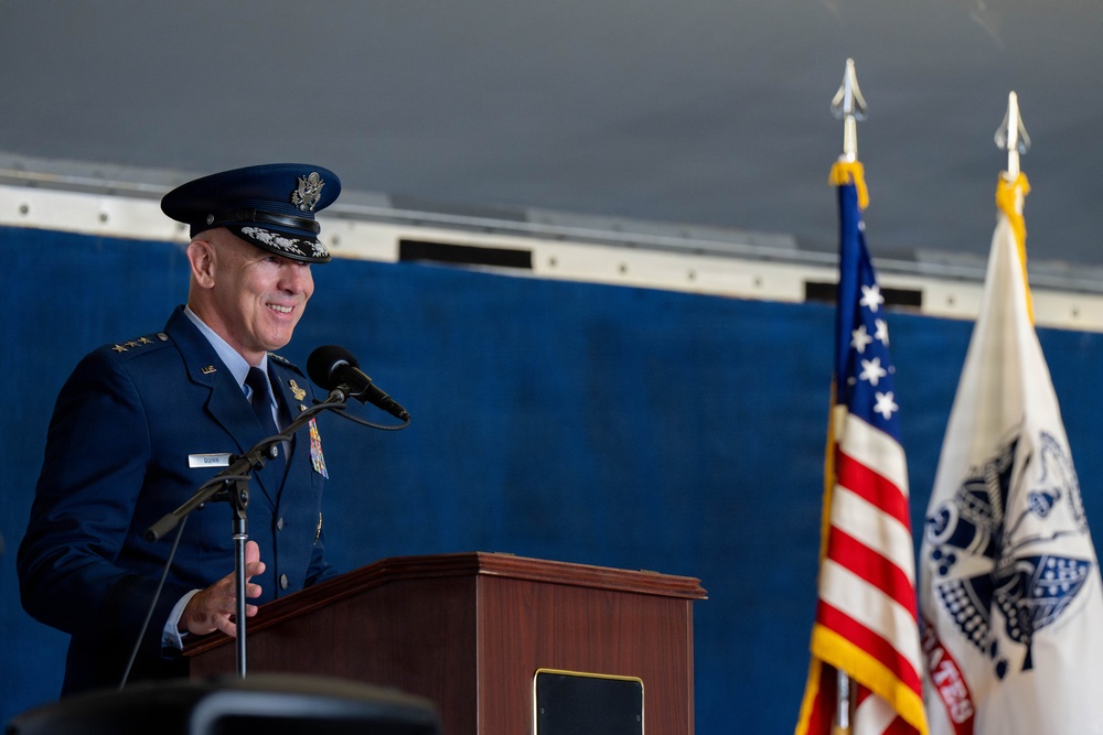 A high-ranking U.S. Air Force officer in dress blues is speaking at a podium during what appears to be a formal military ceremony. The officer wears four-star general insignia and multiple service ribbons, suggesting a significant command-level event. Two flags — the U.S. flag and what appears to be a command or organizational flag — are visible in the background.