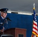 A high-ranking U.S. Air Force officer in dress blues is speaking at a podium during what appears to be a formal military ceremony. The officer wears four-star general insignia and multiple service ribbons, suggesting a significant command-level event. Two flags — the U.S. flag and what appears to be a command or organizational flag — are visible in the background.