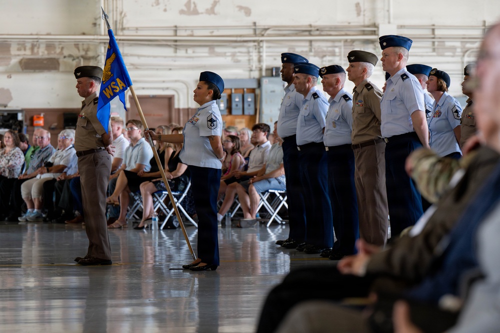 A military ceremony is taking place inside what appears to be a large hangar or drill hall. Personnel in U.S. Air Force dress uniforms and olive/tan service uniforms are standing in formation, with one individual holding a unit guidon flag bearing the letters 'WSA'. Civilian guests and family members are seated in folding chairs observing the ceremony.
