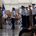 A military ceremony is taking place inside what appears to be a large hangar or drill hall. Personnel in U.S. Air Force dress uniforms and olive/tan service uniforms are standing in formation, with one individual holding a unit guidon flag bearing the letters 'WSA'. Civilian guests and family members are seated in folding chairs observing the ceremony.