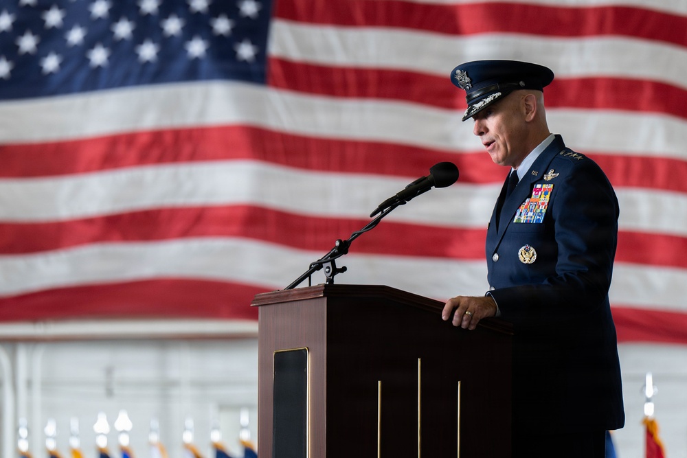 A senior U.S. Air Force officer in dress uniform is delivering a speech at a formal military ceremony from a wooden podium. A large American flag serves as the backdrop, and rows of smaller flags are visible in the background, suggesting a significant official event such as a change of command or retirement ceremony.