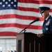A senior U.S. Air Force officer in dress uniform is delivering a speech at a formal military ceremony from a wooden podium. A large American flag serves as the backdrop, and rows of smaller flags are visible in the background, suggesting a significant official event such as a change of command or retirement ceremony.