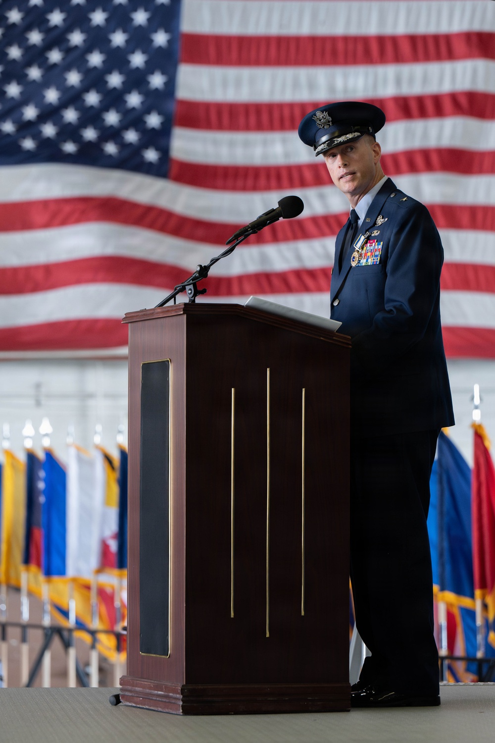 A senior U.S. Air Force officer in dress blues stands at a formal wooden podium delivering a speech at a military ceremony. A large American flag is prominently displayed as a backdrop, and multiple unit guidons or organizational flags are visible in the background. The setting appears to be inside a large hangar or similar military facility.