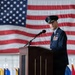 A senior U.S. Air Force officer in dress blues stands at a formal wooden podium delivering a speech at a military ceremony. A large American flag is prominently displayed as a backdrop, and multiple unit guidons or organizational flags are visible in the background. The setting appears to be inside a large hangar or similar military facility.