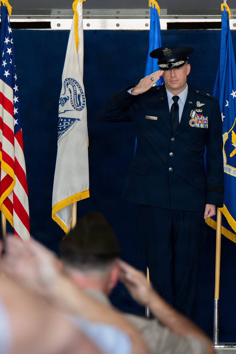 A senior U.S. Air Force officer in dress blues is rendering a salute on a ceremonial stage, likely during a change of command or activation ceremony. The officer wears a service cap with general/senior officer oak leaf scramble, pilot wings, and multiple rows of service ribbons. Audience members in the foreground are also saluting in return.