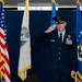 A senior U.S. Air Force officer in dress blues is rendering a salute on a ceremonial stage, likely during a change of command or activation ceremony. The officer wears a service cap with general/senior officer oak leaf scramble, pilot wings, and multiple rows of service ribbons. Audience members in the foreground are also saluting in return.